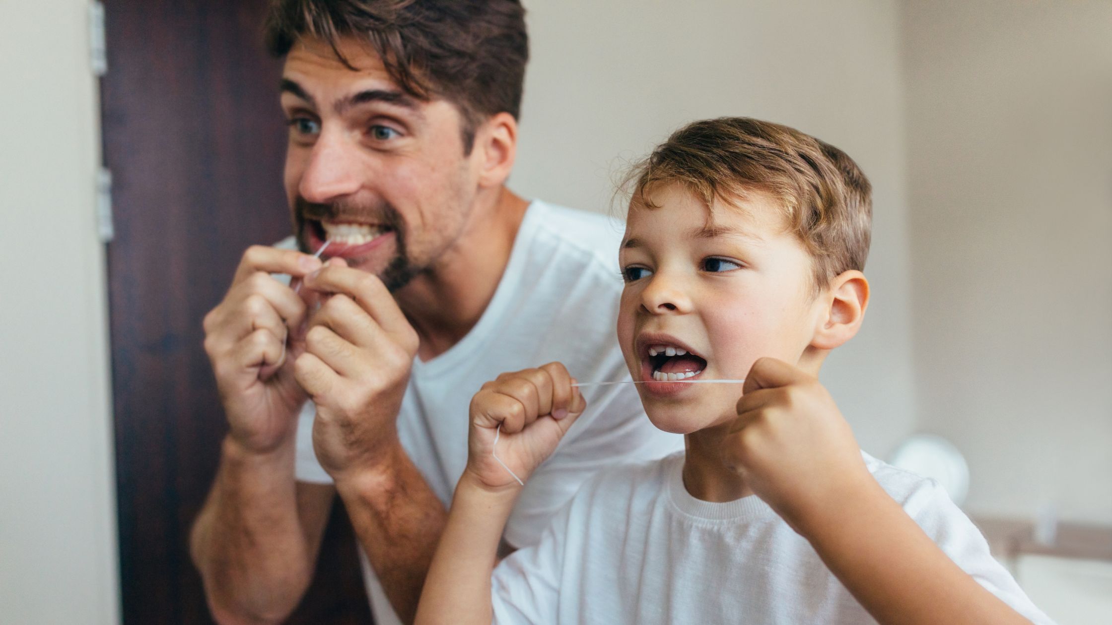 Father and son flossing side by side in the bathroom.